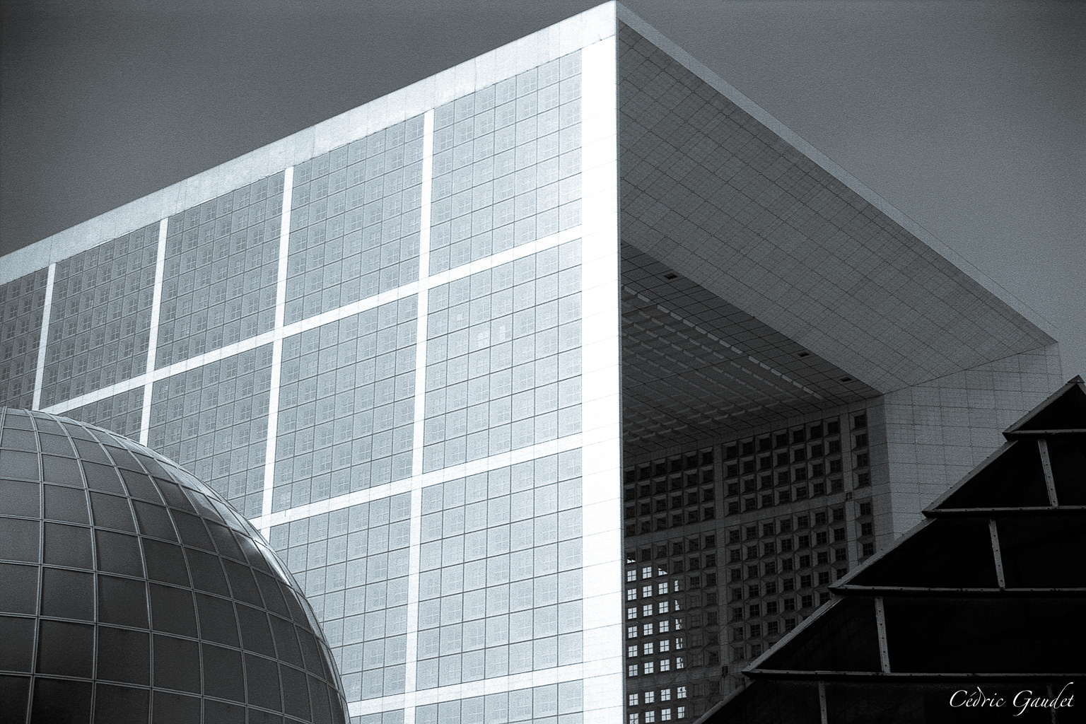 Vue en gros plan d'une façade architecturale moderne, mélange de béton, verre et acier, avec un angle marqué pointant vers le ciel. En arrière-plan, une sphère en verre et une pyramide de verre sont visibles, créant une composition intéressante.