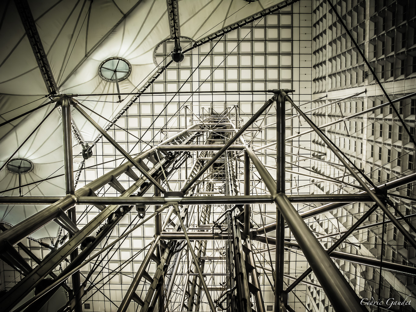 Vue en contre-plongée d'une structure architecturale complexe composée de béton, d'acier et de verre, avec des câbles et des supports interconnectés, sous un plafond lumineux.
