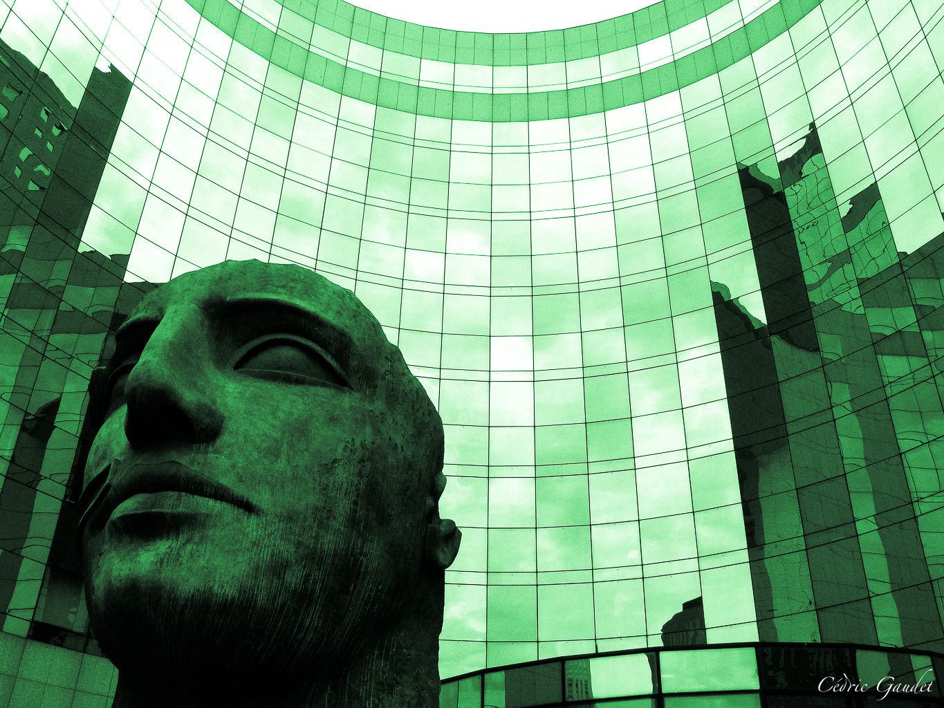 Statue en gros plan d'un visage stylisé, avec un fond de verres et de reflets de bâtiments modernes.